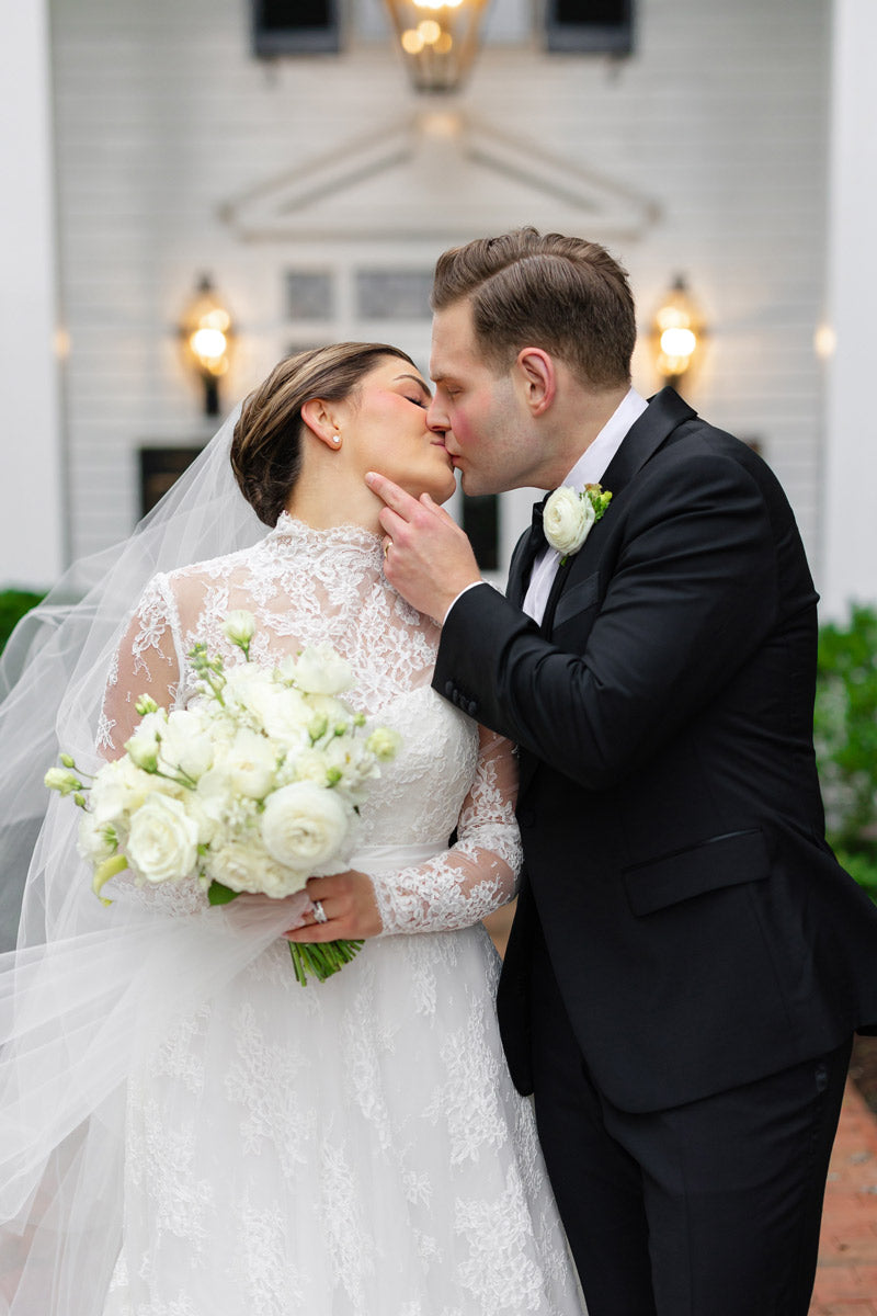 A bride and groom share a kiss outside, dressed in a lace wedding gown and a black suit. The bride holds a white bouquet, and both wear white boutonnieres, standing in front of a white building.