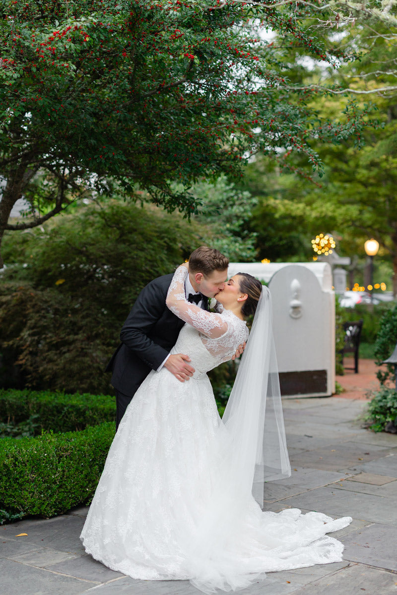 A groom in a black suit gently dips and kisses his bride, who wears a long-sleeved white lace gown and veil, in a lush garden setting with greenery and twinkling lights in the background.