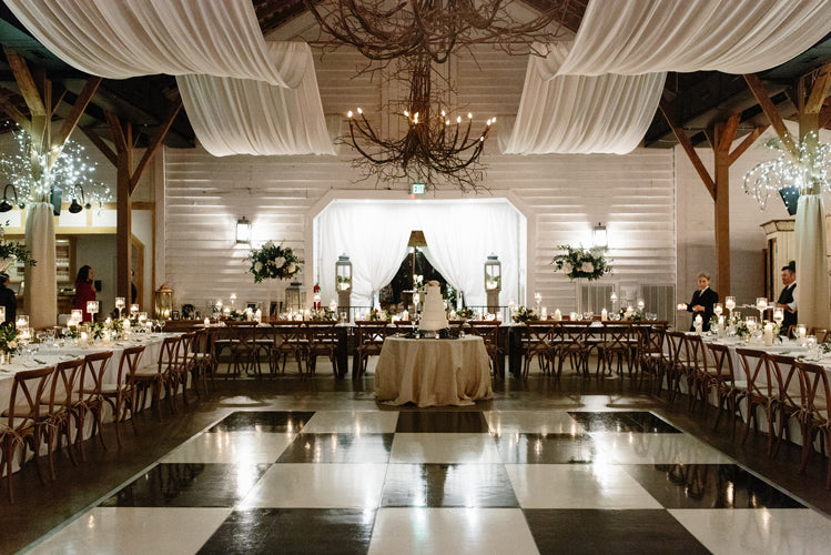 A decorated wedding reception hall with draped white fabric on the ceiling, chandeliers, long banquet tables adorned with candles and flowers, and a round table with a tiered cake at the center, on a black-and-white checkered floor.