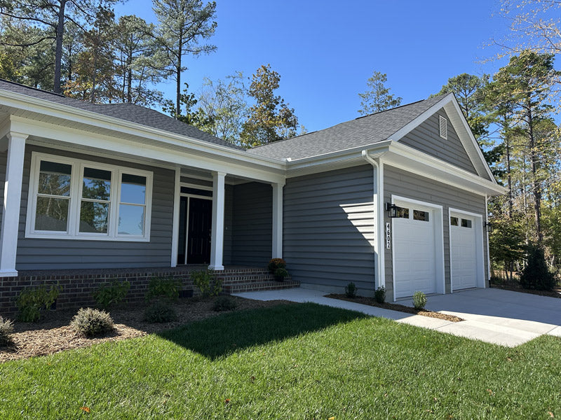 A modern single-story house with gray siding, white trim, a black front door, and two white garage doors. The home has a small porch, neatly trimmed grass, and landscaped bushes in front. Tall trees are in the background under a clear sky.