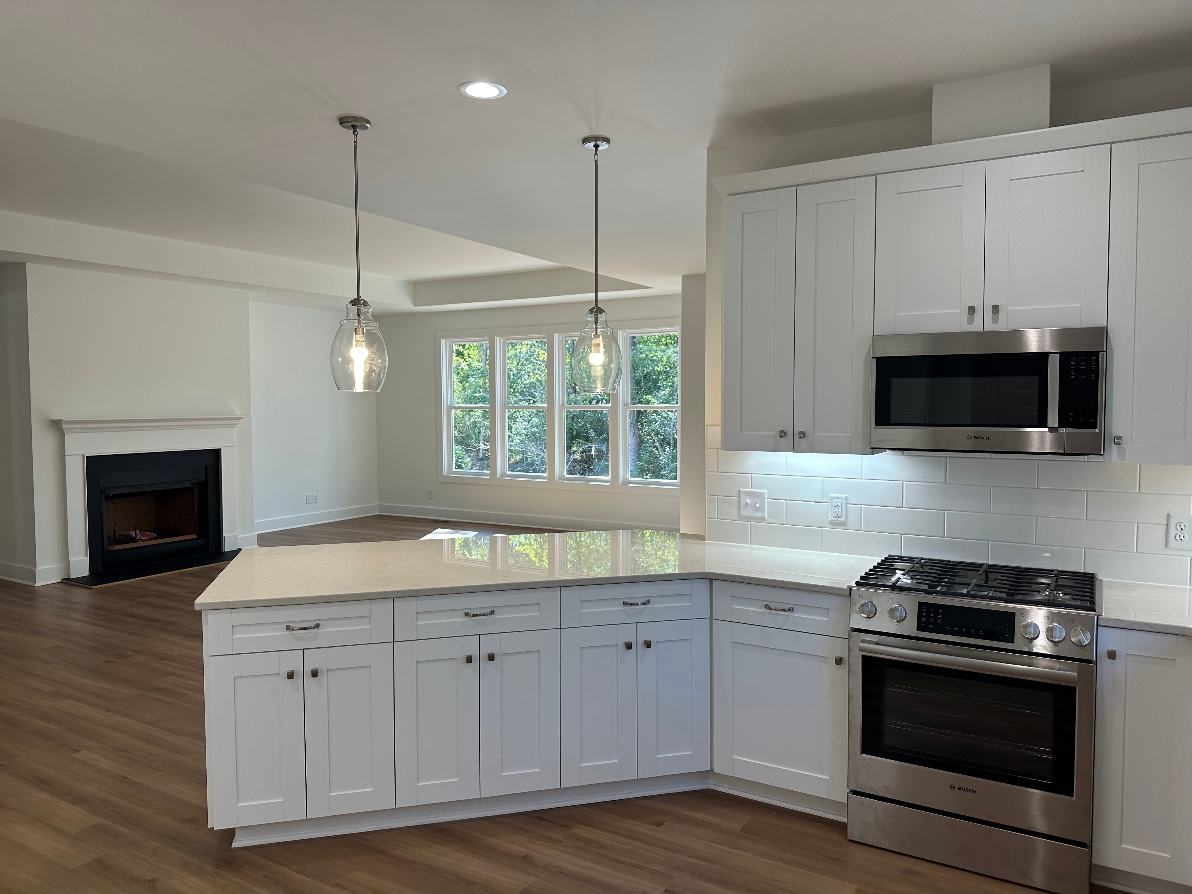 Bright, modern kitchen with white cabinets, stainless steel appliances, a gas range, and subway tile backsplash. Two pendant lights hang over a peninsula counter. Open living area with fireplace and large windows in the background.