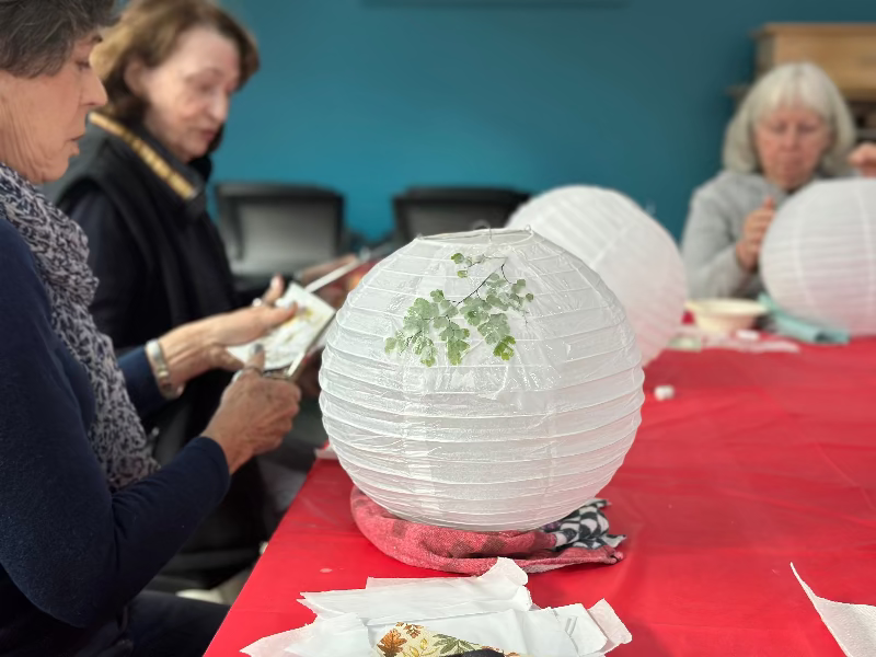 Three women sit around a red table working on white paper lanterns, decorating them with leaves and botanical designs. The focus is on a lantern in the foreground with a green leaf glued to it.
