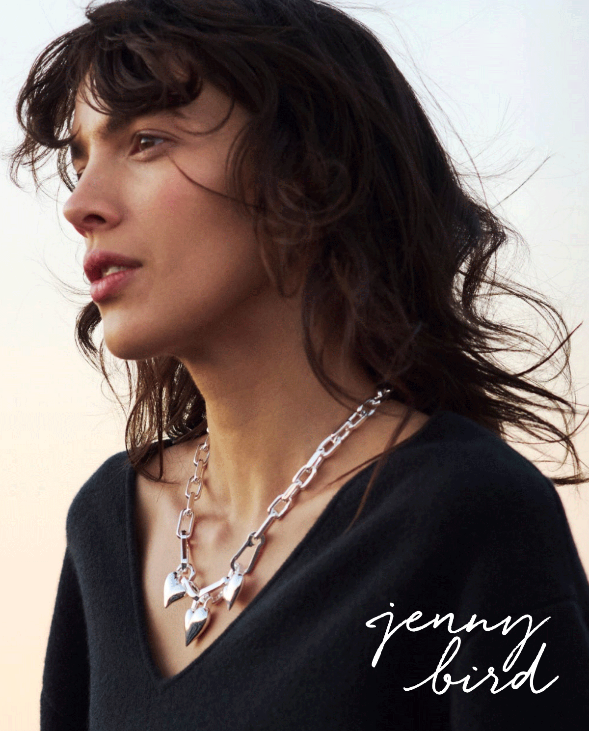 A woman with wavy dark hair wears a chunky silver chain necklace and a black V-neck top, looking into the distance. The text “jenny bird” appears in cursive at the bottom right. The background is softly lit.