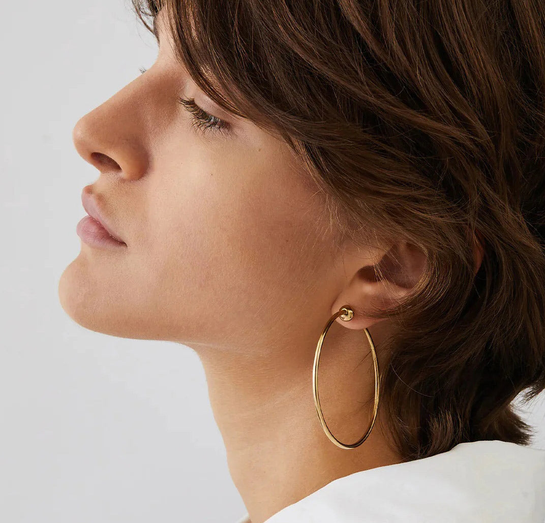 A person with short brown hair is shown in profile facing left, wearing a large gold hoop earring. Their eyes are closed and the background is plain and light-colored.