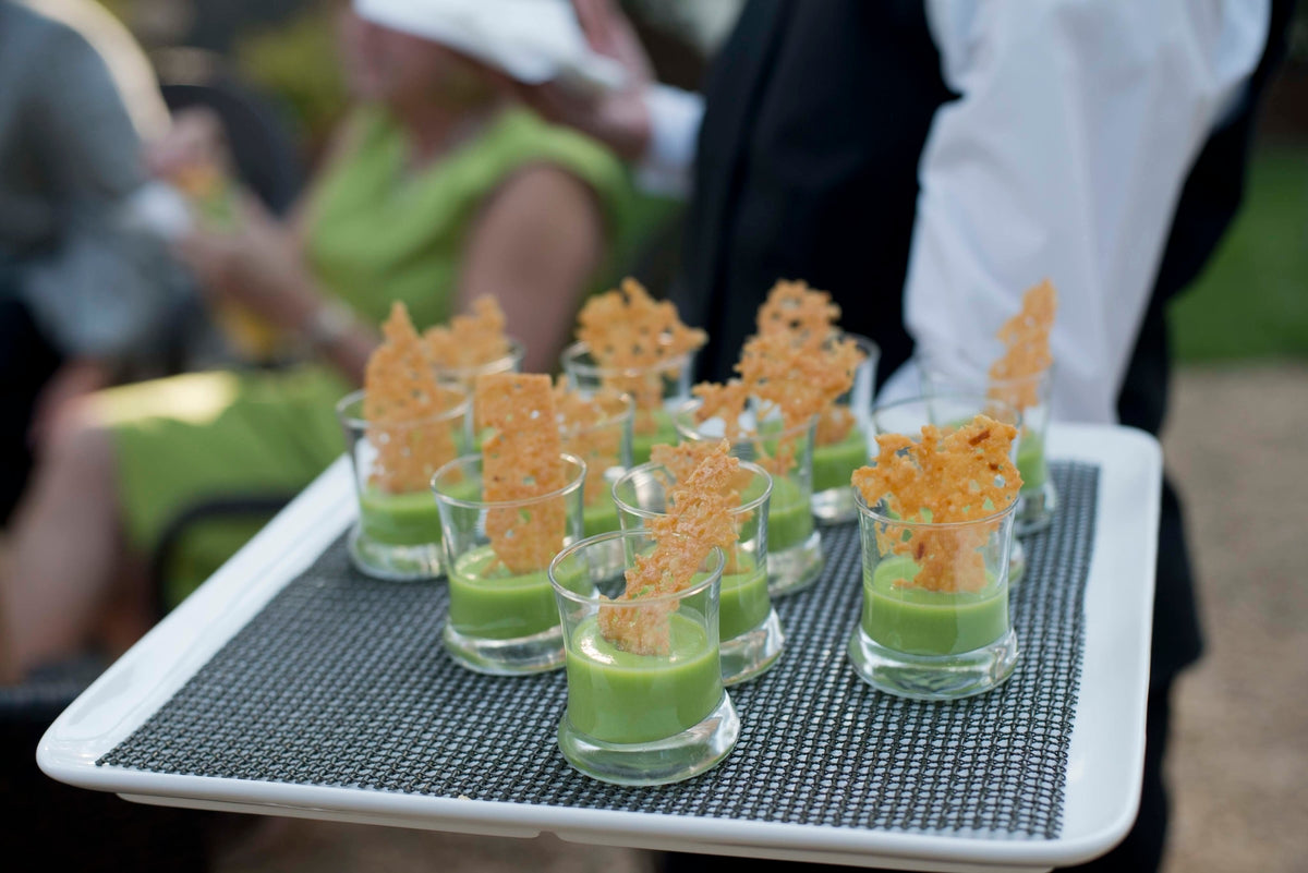A server holds a tray of small glasses filled with green soup, each topped with a crisp cheese wafer. The tray is being carried at what appears to be an outdoor event, with blurred guests in the background.