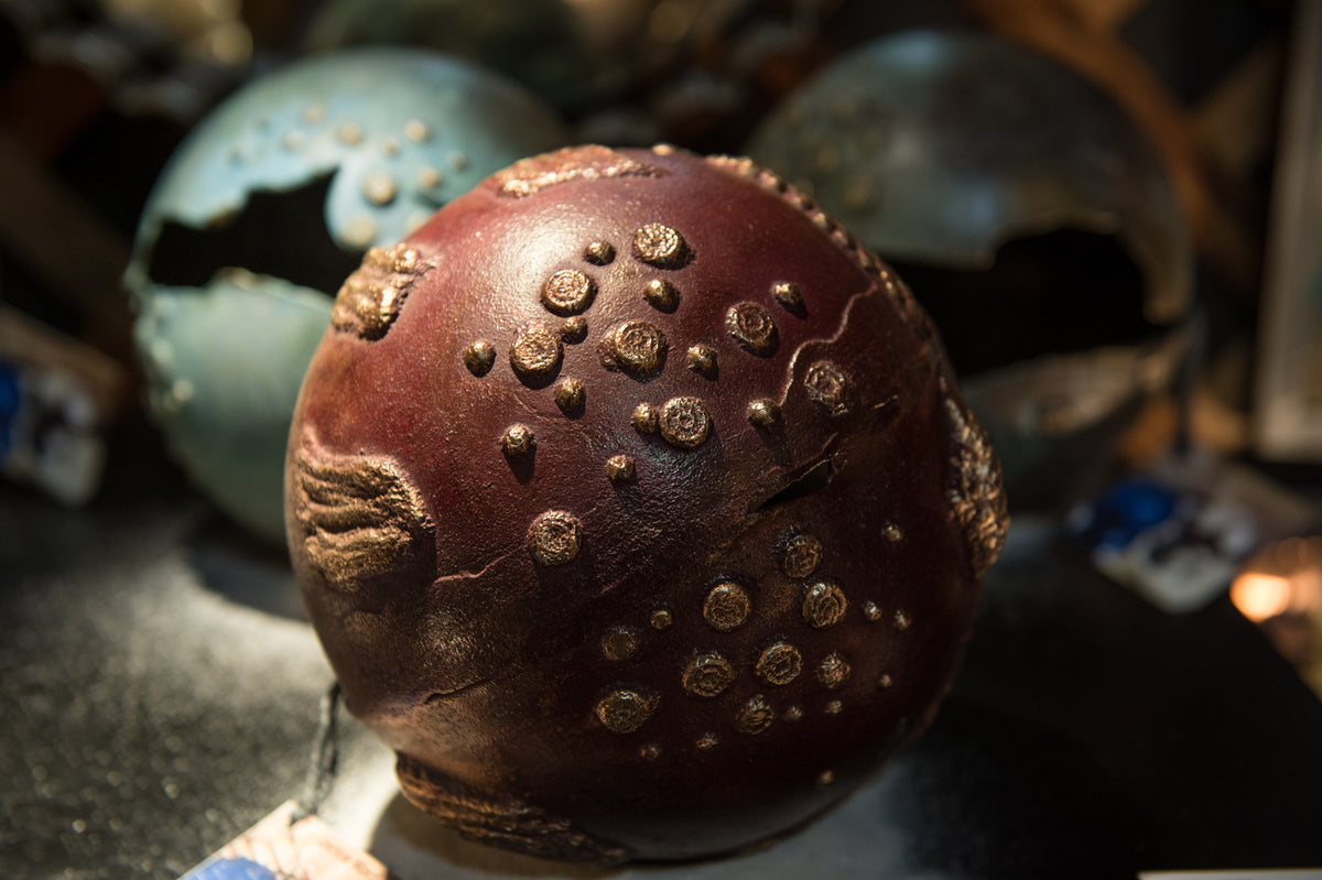 A close-up of a spherical, reddish-brown object with raised, circular bumps and textured patches, resembling a fantasy or alien artifact. Two similar, greenish spheres are blurred in the background.