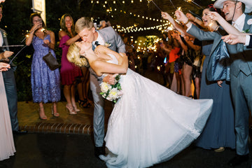 A newlywed couple shares a romantic kiss as the groom dips the bride. They are surrounded by guests in formal attire holding sparklers under string lights, celebrating their wedding at night.