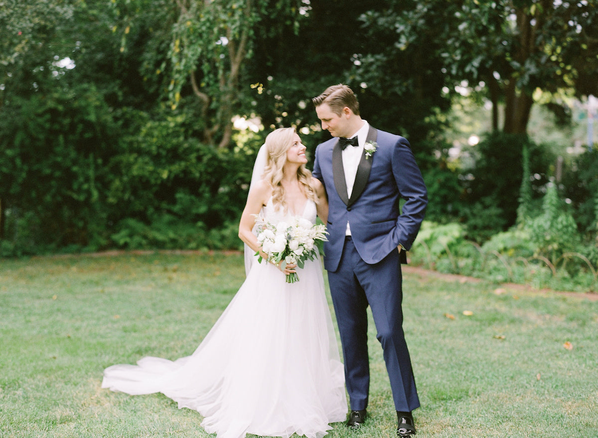 A bride in a white gown and a groom in a blue suit stand together on grass, smiling at each other, surrounded by lush green trees and plants.