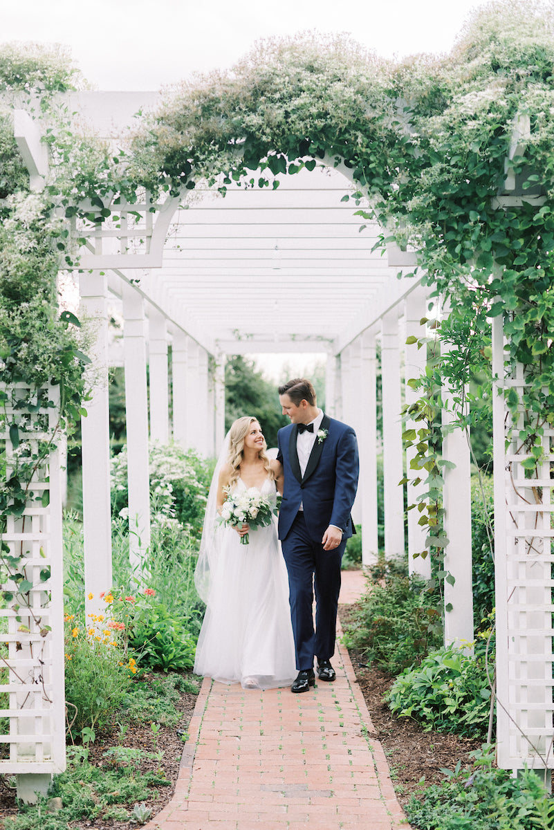 A bride and groom walk arm in arm under a white pergola covered with greenery and flowers. The bride holds a bouquet and wears a veil, while the groom is in a navy suit and bow tie. They smile at each other in a garden setting.