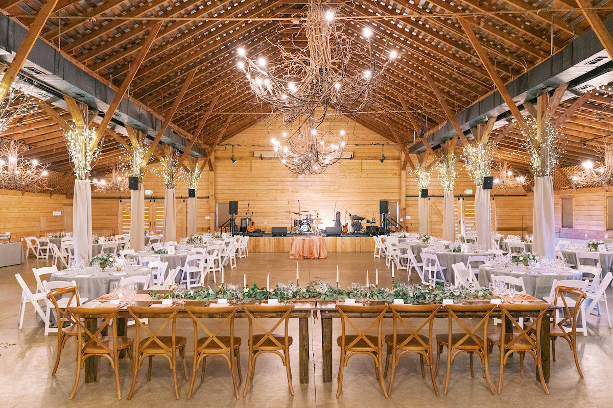Rustic wedding reception in a wooden barn with string lights, tables with white chairs, greenery centerpieces, a head table in front, and a stage with musical instruments at the back.