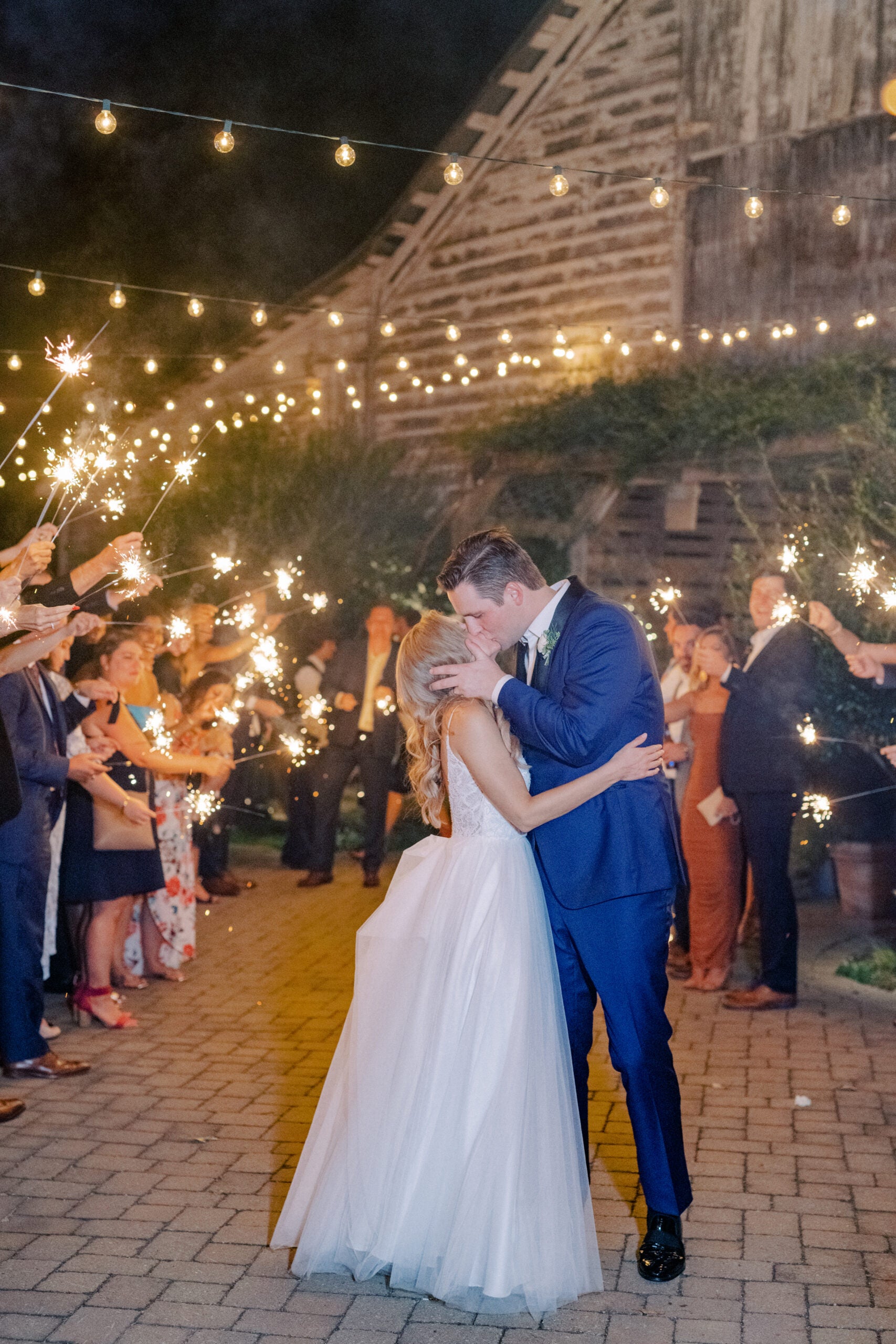 A bride and groom share a kiss outside at night under string lights, surrounded by guests holding sparklers. The scene is festive, with a rustic barn in the background and people celebrating their wedding.