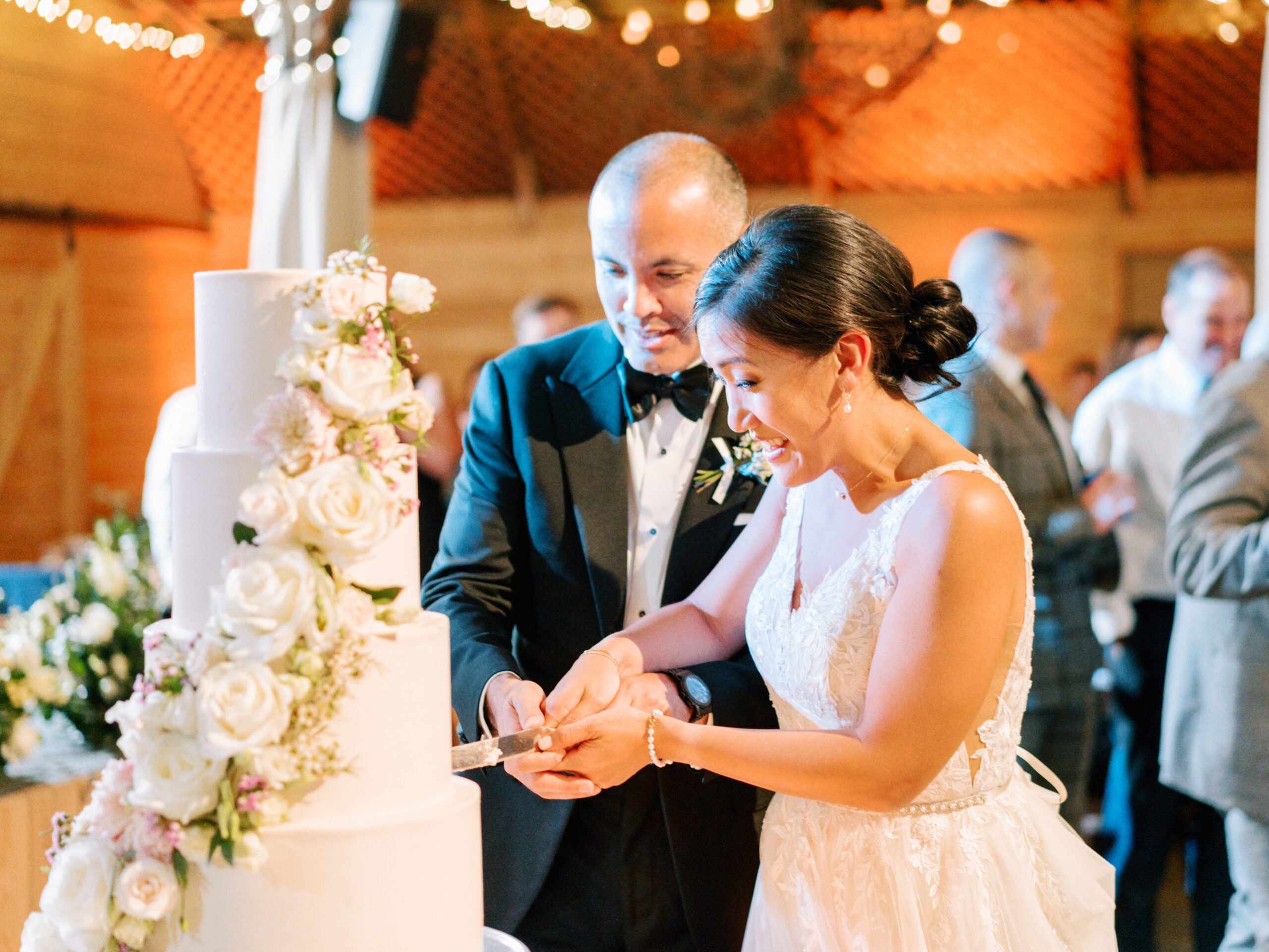 A bride and groom, both smiling, cut a tall white wedding cake decorated with white roses and flowers at their reception, surrounded by warmly lit guests in the background.