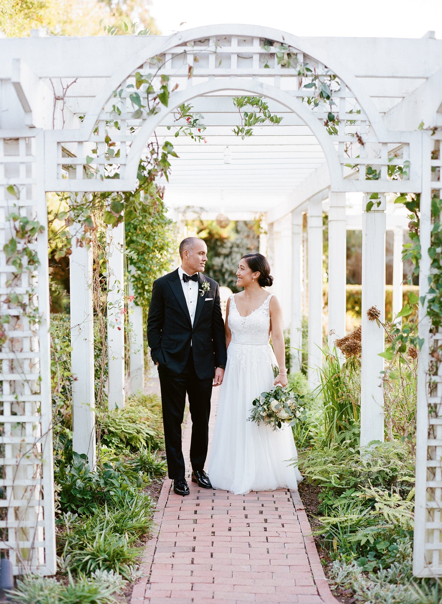 A bride and groom walk hand in hand under a white garden arbor, surrounded by greenery. The groom wears a black tuxedo, and the bride wears a white dress and holds a bouquet of flowers. Both are smiling at each other.