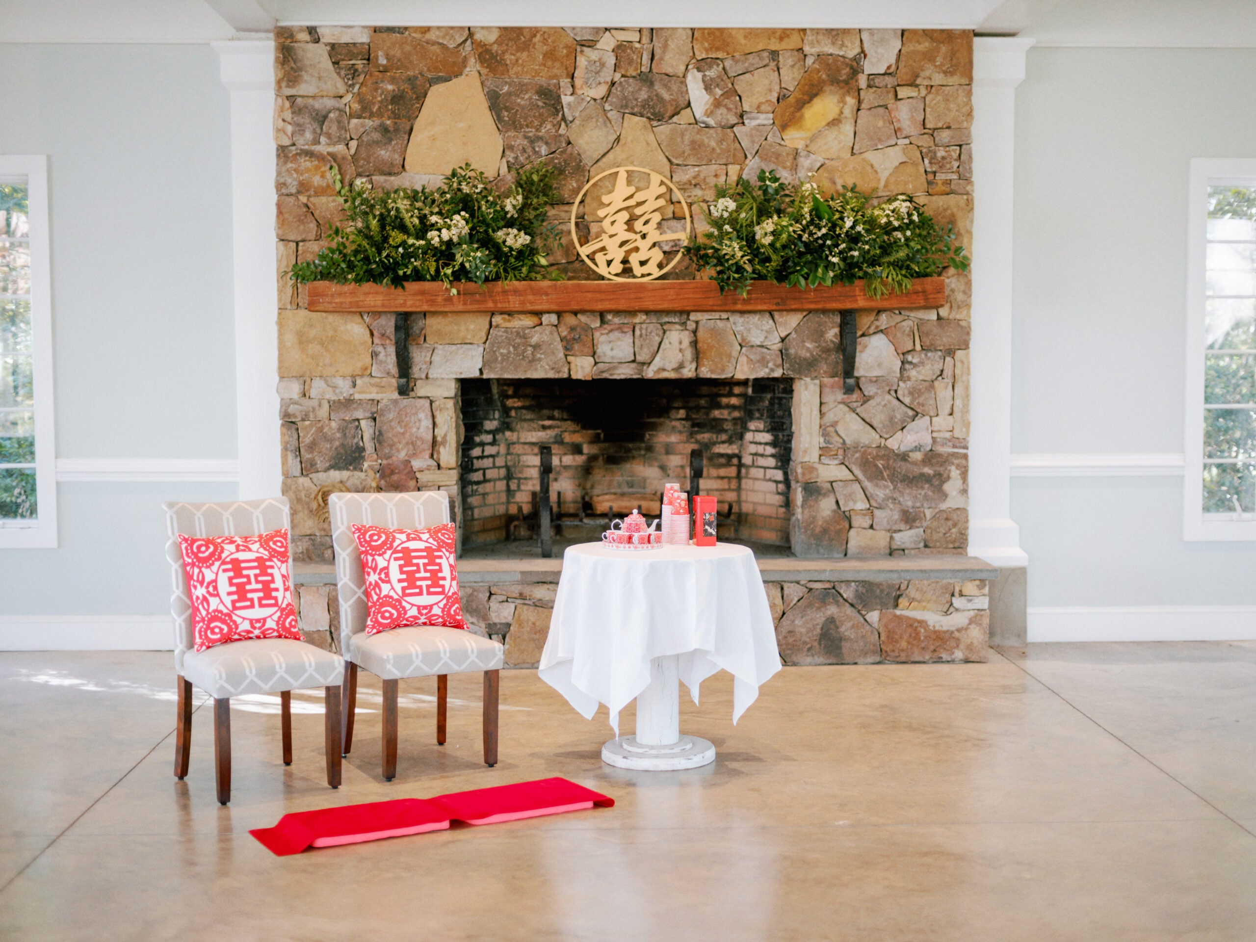 A stone fireplace is decorated with greenery and a large gold double happiness symbol. In front, two chairs with red double happiness pillows and a table with tea items are arranged for a traditional tea ceremony.
