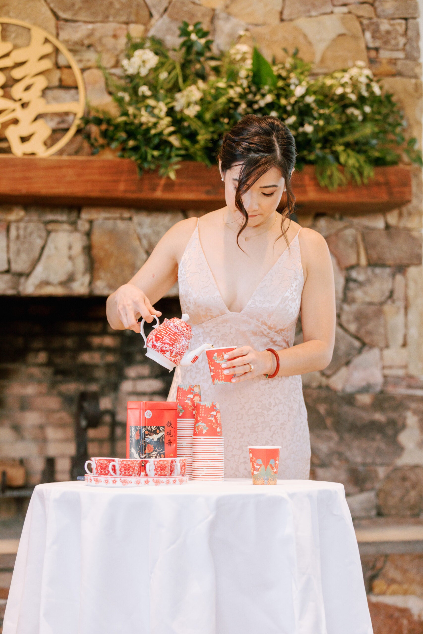 A woman in a light dress pours tea from a red teapot into a matching cup at a table with red teacups and a white cloth, set in front of a stone fireplace with greenery and a wooden decoration.