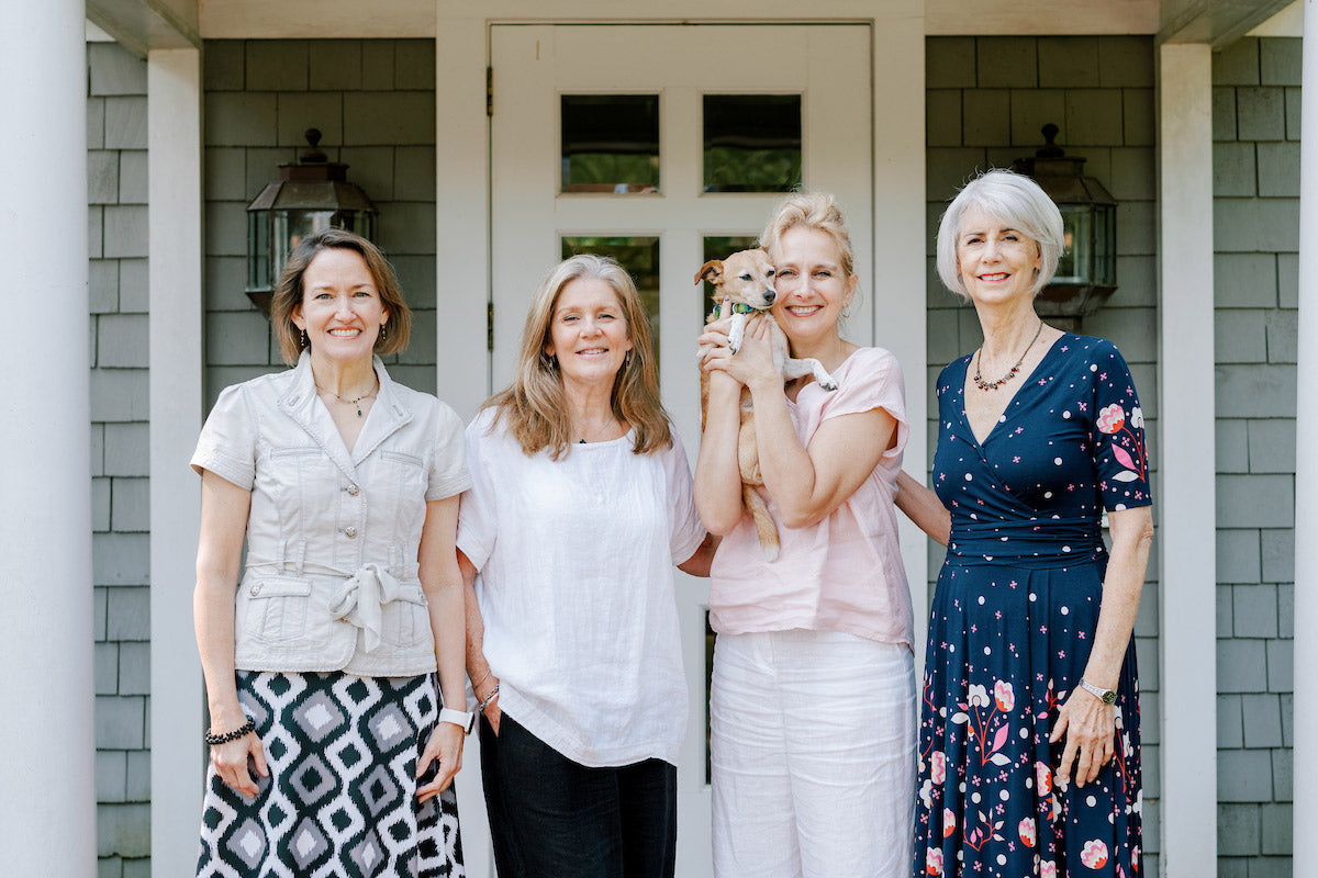 Four women stand smiling in front of a house entrance; one woman is holding a small dog. The group appears happy and casually dressed, posing for a photo on the front porch.