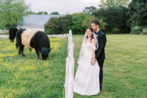 A bride and groom stand together in a grassy field near a white fence, smiling. Behind them, a black and white cow grazes. Trees and a building are visible in the background.