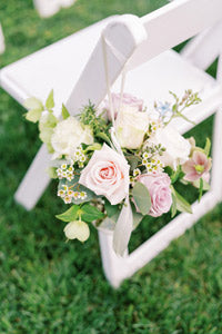 A small floral arrangement with pastel roses and greenery hangs from a white ribbon on the back of a white folding chair set on green grass.