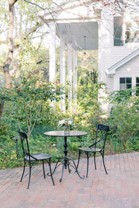 A small round table with two black metal chairs sits on a brick patio. A vase with flowers is on the table. Lush greenery and a white house with columns are in the background.