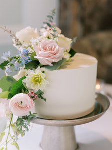 A simple white cake decorated with pink and white roses, greenery, and small blue flowers, displayed on a silver cake stand.