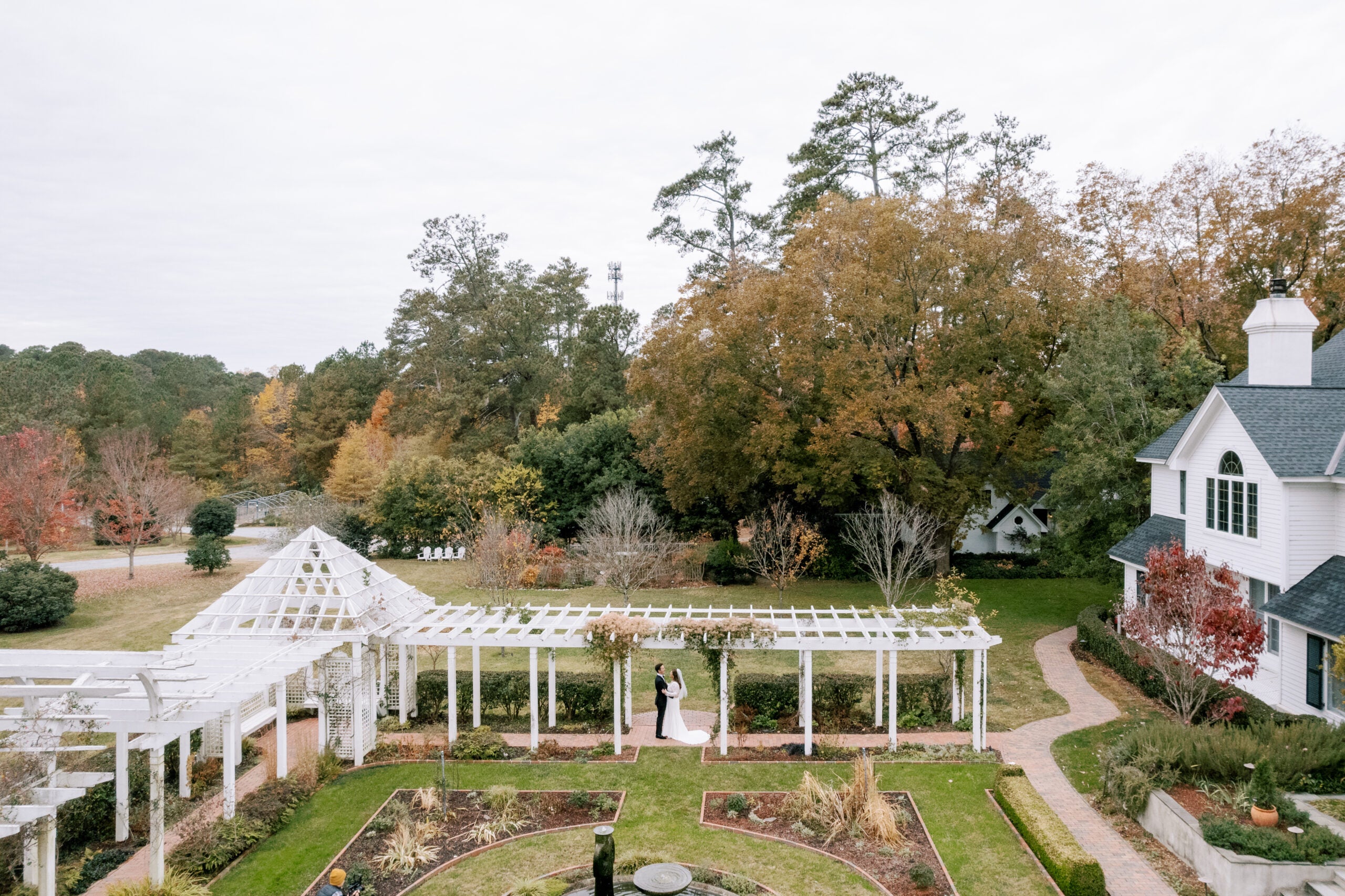 A bride and groom stand under a white pergola in a formal garden, surrounded by autumn trees and greenery, next to a white house with a dark roof. The scene is tranquil and romantic.