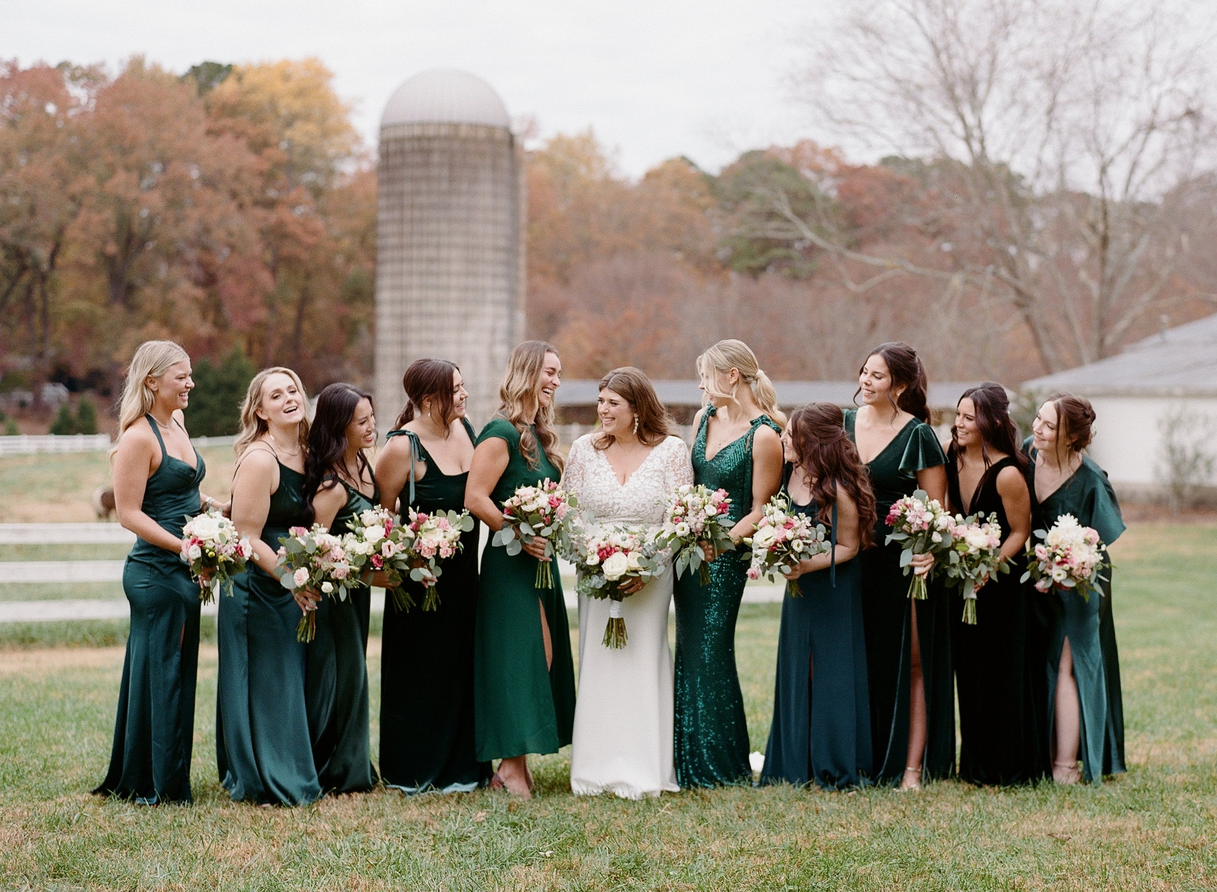 A bride in a white dress stands outdoors with ten bridesmaids in dark green dresses, all holding bouquets and smiling. The group is on grass, with autumn trees, a silo, and a white fence in the background.