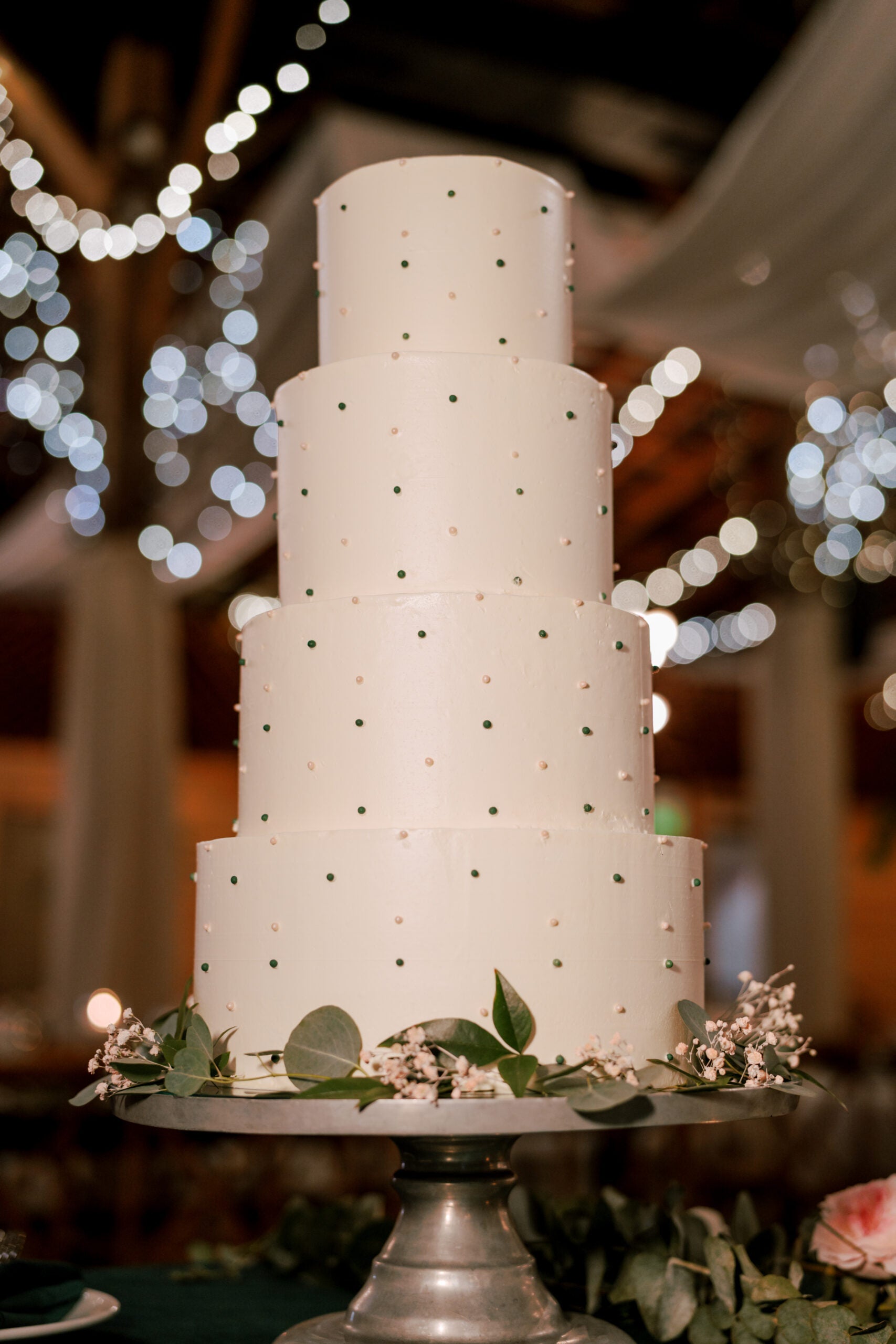 A tall, five-tiered white wedding cake decorated with small green and white dots, topped with greenery and white flowers, sits on a silver stand. Blurred string lights twinkle in the background.