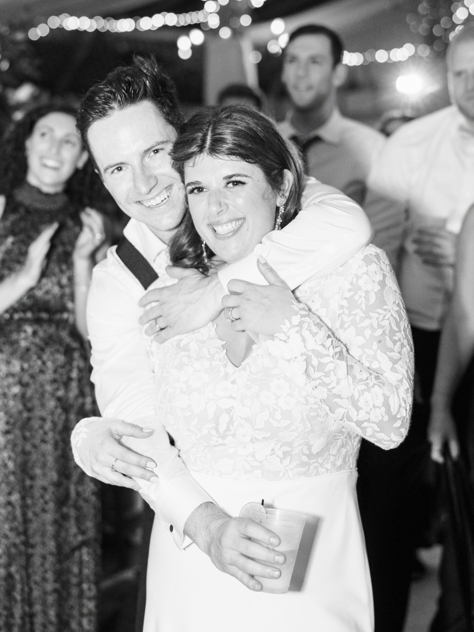 A joyful couple embraces and smiles at the camera during a celebration, with guests clapping and string lights twinkling in the background. The woman wears a white lace dress and holds a cup. The image is in black and white.