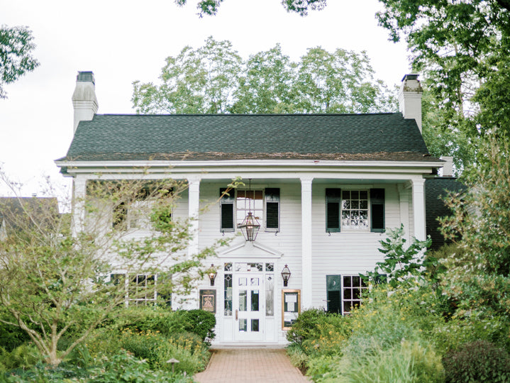 A charming two-story white house with black shutters, a dark roof, and two chimneys. The front has columns and a glass door, surrounded by lush green trees and bushes along a brick walkway.