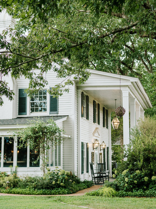 A classic white house with black shutters and columns sits surrounded by lush greenery and trees. Two black rocking chairs and hanging lanterns are on the front porch, creating a peaceful, inviting atmosphere.