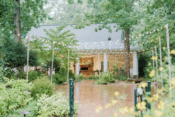 A lush garden with string lights hanging above a brick pathway leads to a house with white pillars. Green trees and plants surround the area, creating a serene and festive outdoor setting.
