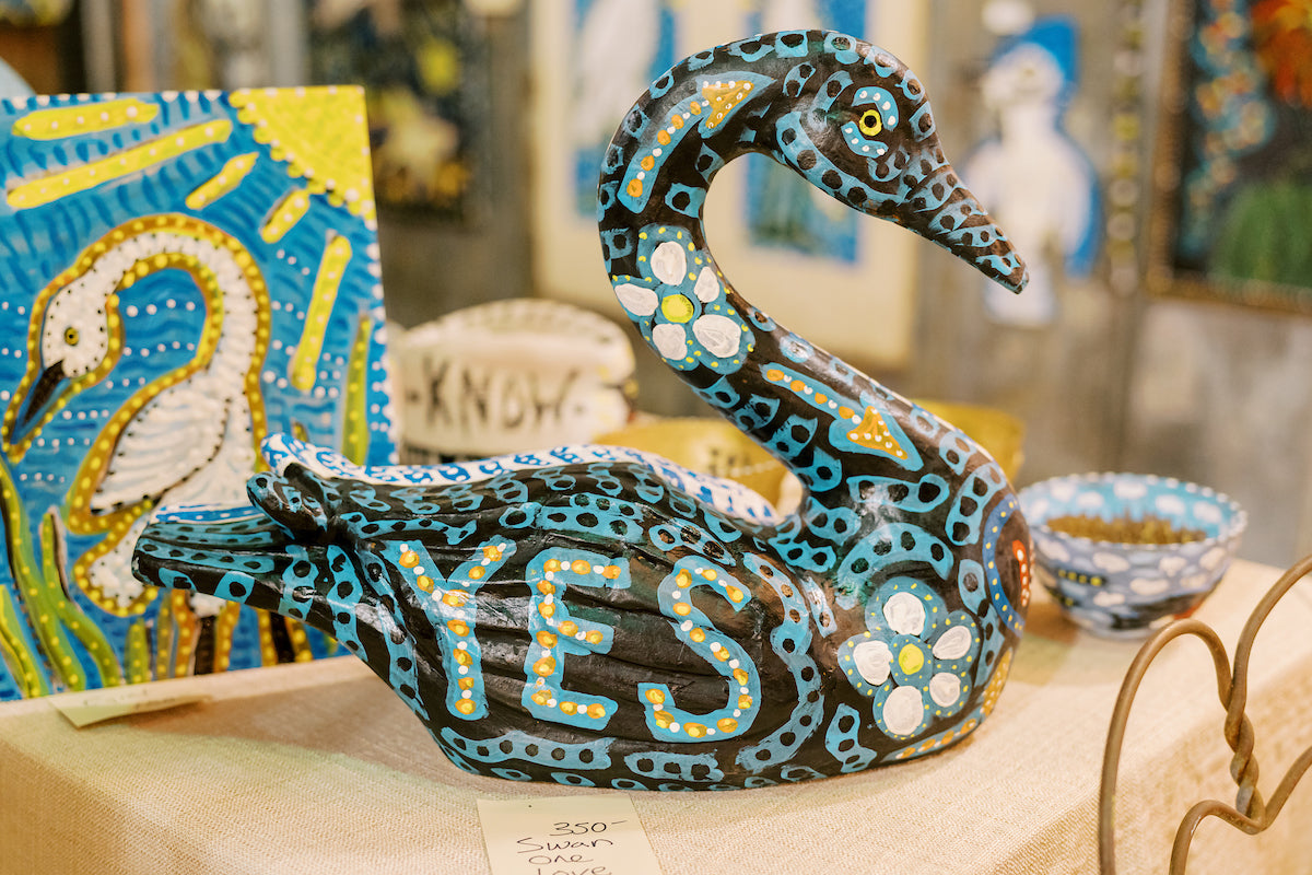 A painted sculpture of a black swan with blue, yellow, and white dots and patterns, featuring the word "YES" on its side, displayed on a table with other colorful artwork in the background.