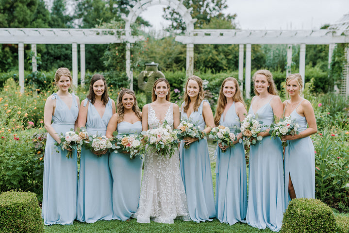 A bride in a white lace dress stands outdoors with seven bridesmaids in light blue dresses, all holding bouquets, in a lush garden with white trellises and greenery in the background.