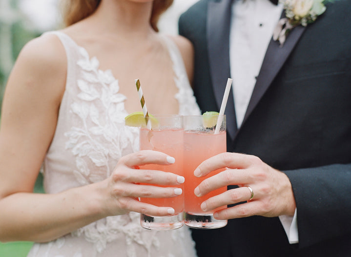 A bride and groom in formal attire hold pink cocktails with lime garnishes and straws, standing close together. The bride wears a white dress with floral lace, and the groom wears a black tuxedo with a boutonniere.