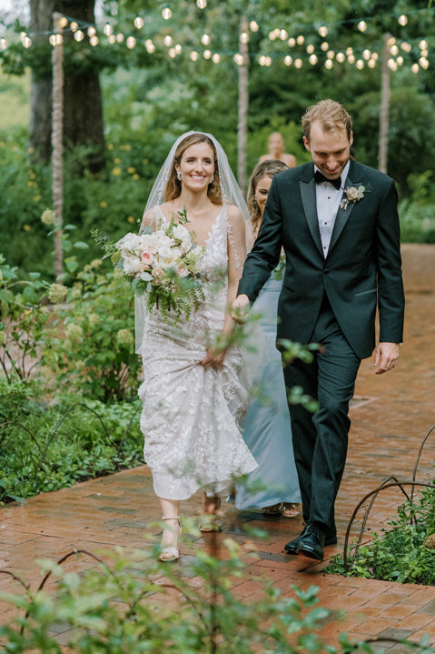 A bride in a white dress and veil holds a bouquet while walking with a groom in a black tuxedo along a brick path, surrounded by greenery and string lights. Both are smiling as they walk outdoors.