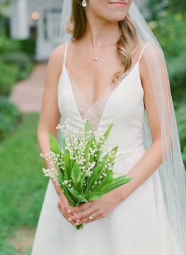 A bride stands outdoors in a white dress with a deep V neckline and sheer details, holding a bouquet of lily of the valley flowers. She wears a veil and simple jewelry, with greenery and a walkway in the background.