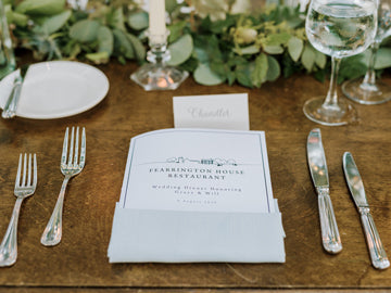 A formal place setting on a wooden table features a menu tucked into a napkin, labeled