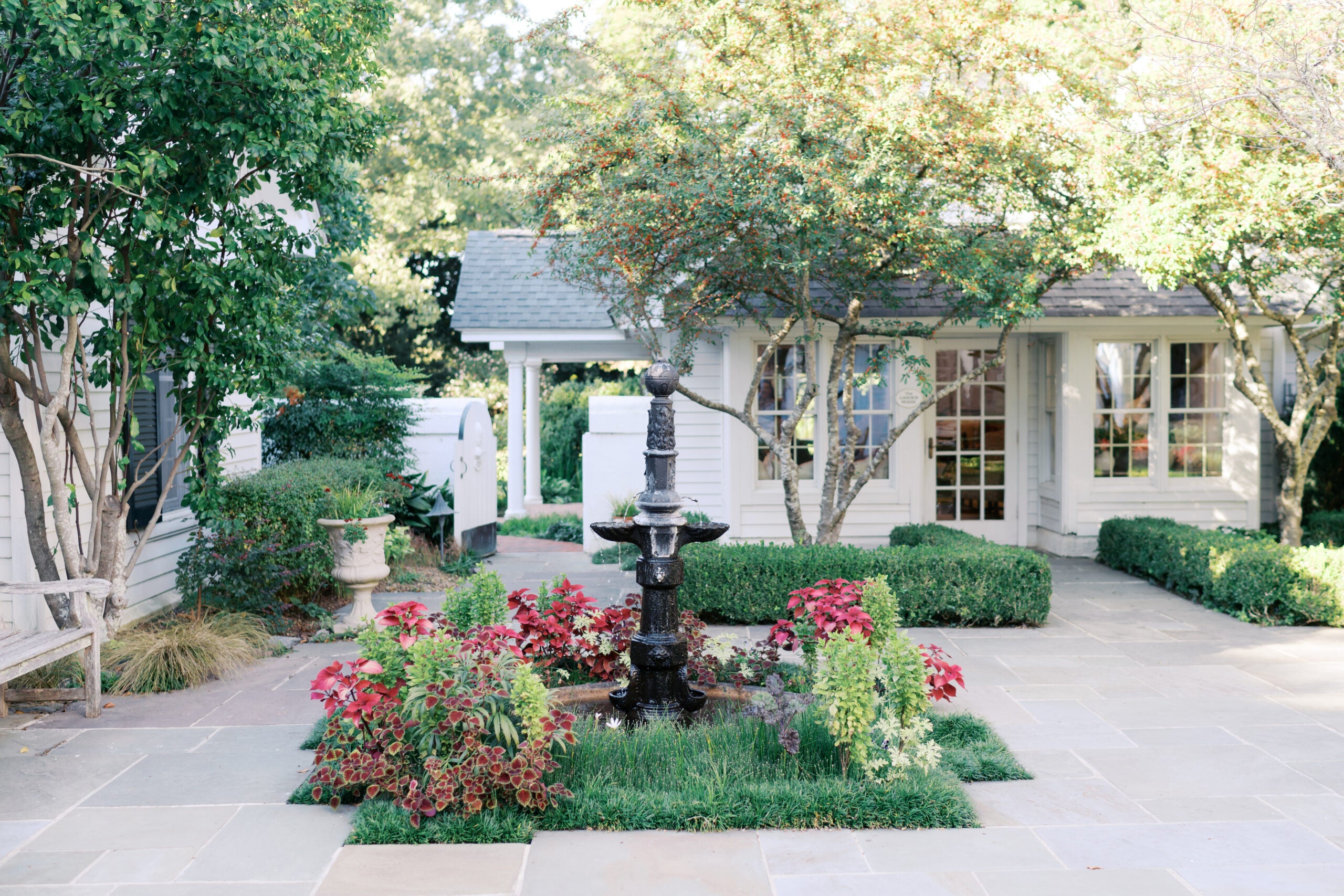 A small black fountain surrounded by red and green plants sits in a paved courtyard with manicured bushes, trees, and a white house with large windows in the background.