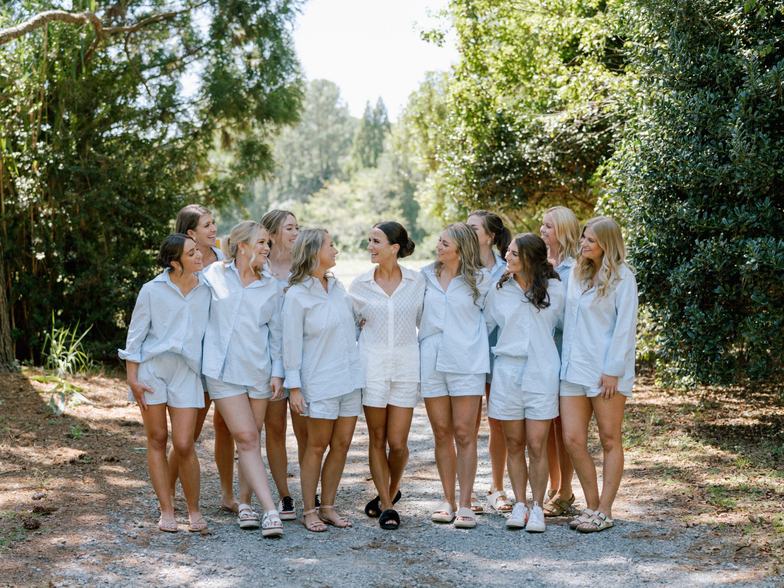 A group of ten women, all wearing light-colored matching outfits, stand outdoors on a tree-lined gravel path, smiling and looking at each other in a relaxed, cheerful atmosphere.