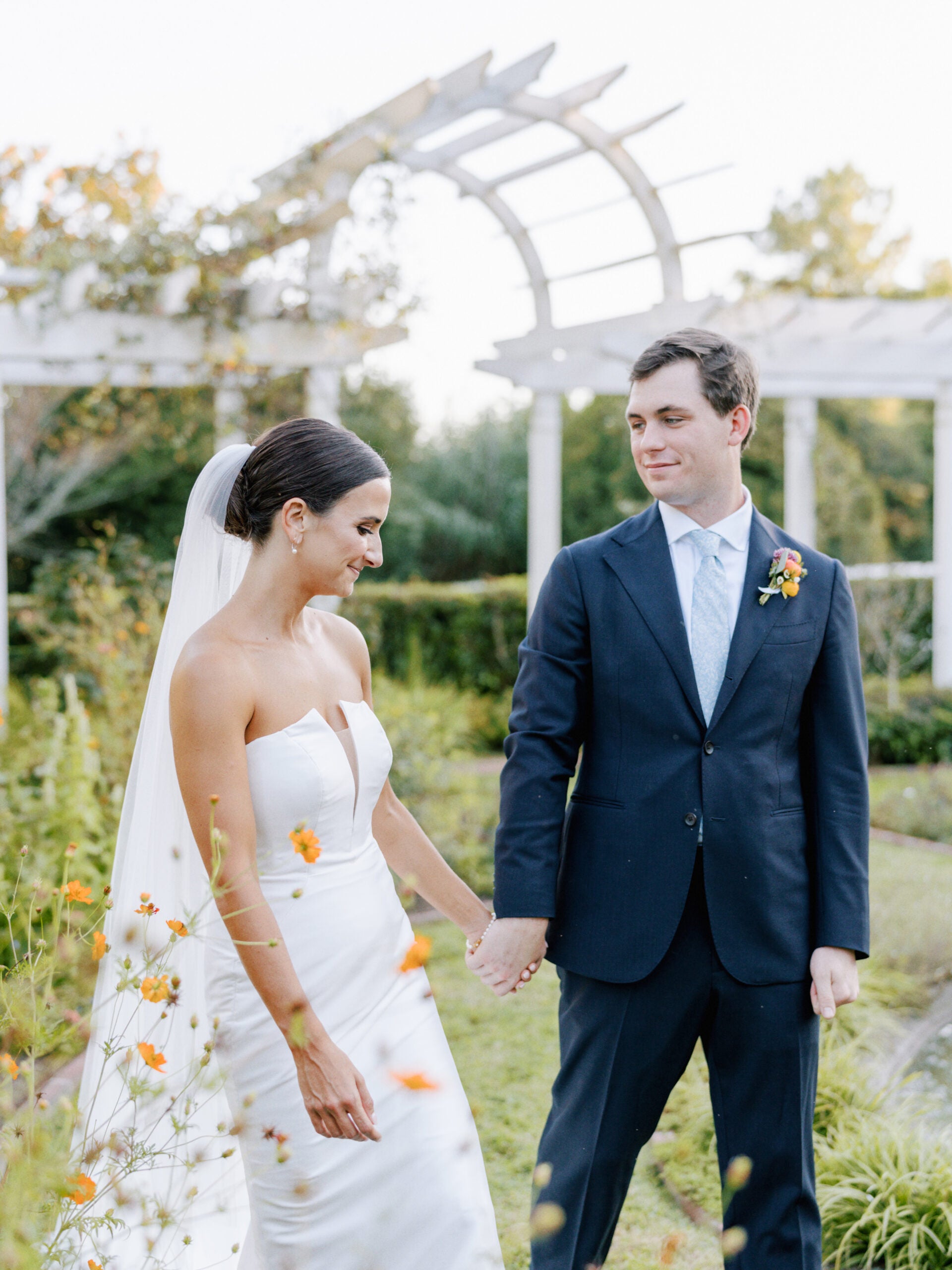 A bride in a strapless white gown and veil holds hands with a groom in a navy suit and blue tie, standing together outdoors in a garden with orange flowers and a white arbor in the background.