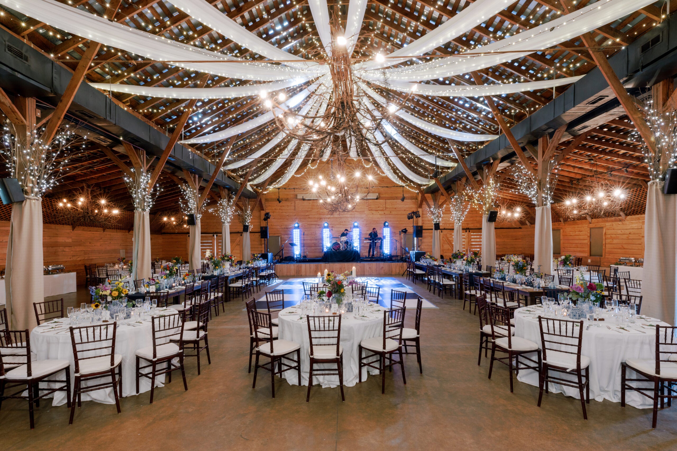 A rustic barn venue decorated for an event with round tables, white tablecloths, floral centerpieces, string lights, draped fabric on the ceiling, and a stage with instruments at the far end.