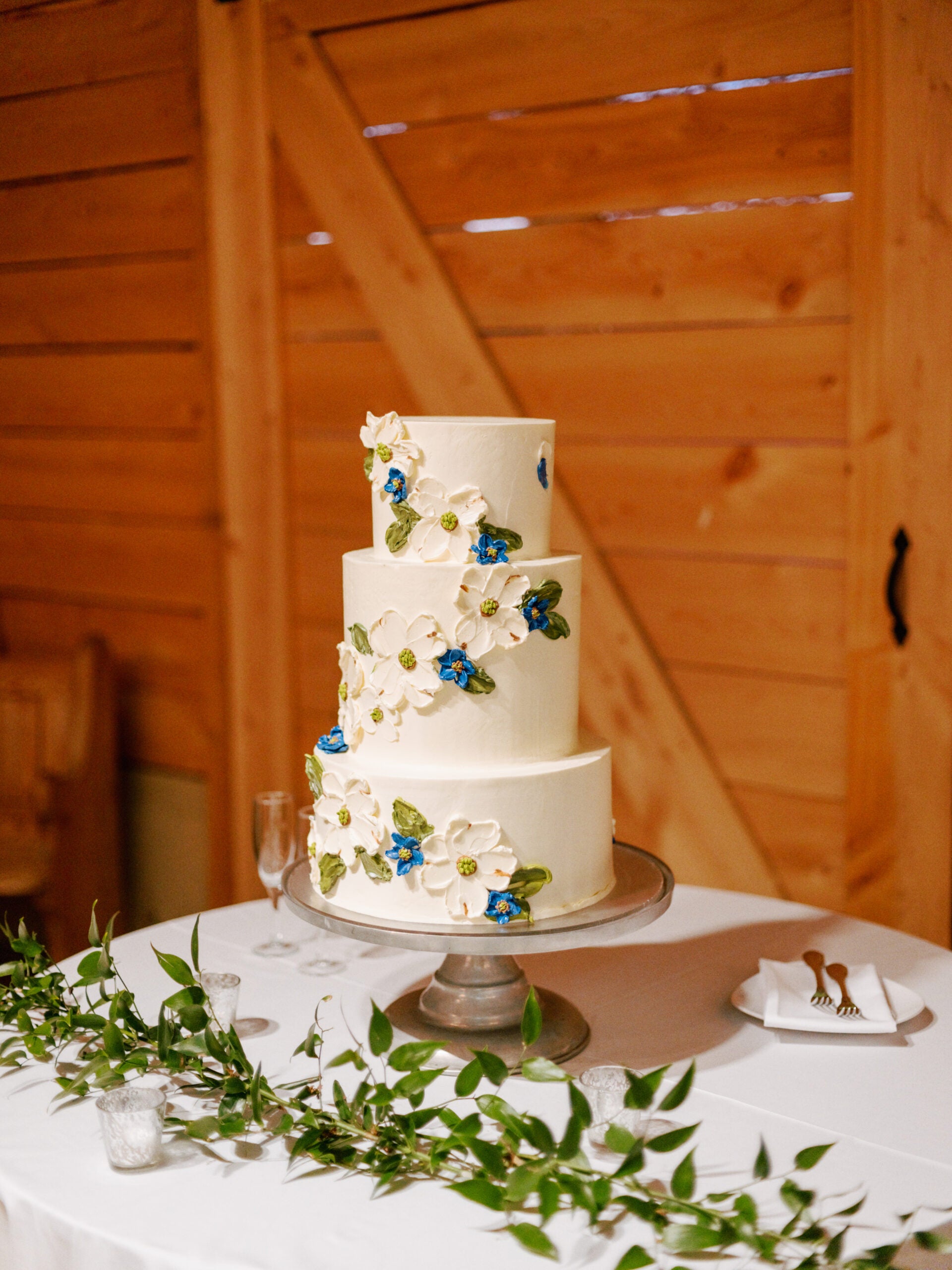 A three-tiered white wedding cake decorated with large white and blue flowers sits on a silver cake stand on a round table, with green leaves and candles as decoration. The background features wooden walls and a barn door.