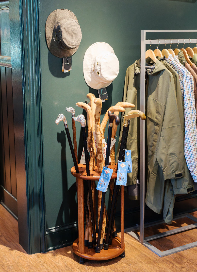 A display in a store shows wooden walking canes in a stand, hats hanging on a green wall, and a clothing rack with jackets and shirts on wooden hangers.