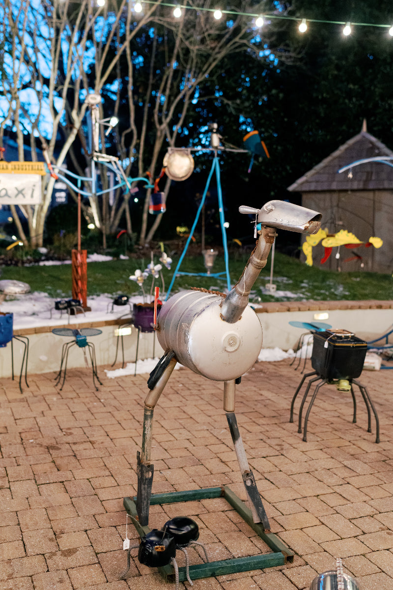 A metal bird sculpture made from recycled parts stands on a brick patio at night, surrounded by other whimsical metal sculptures under string lights, with trees and a small shed in the background.