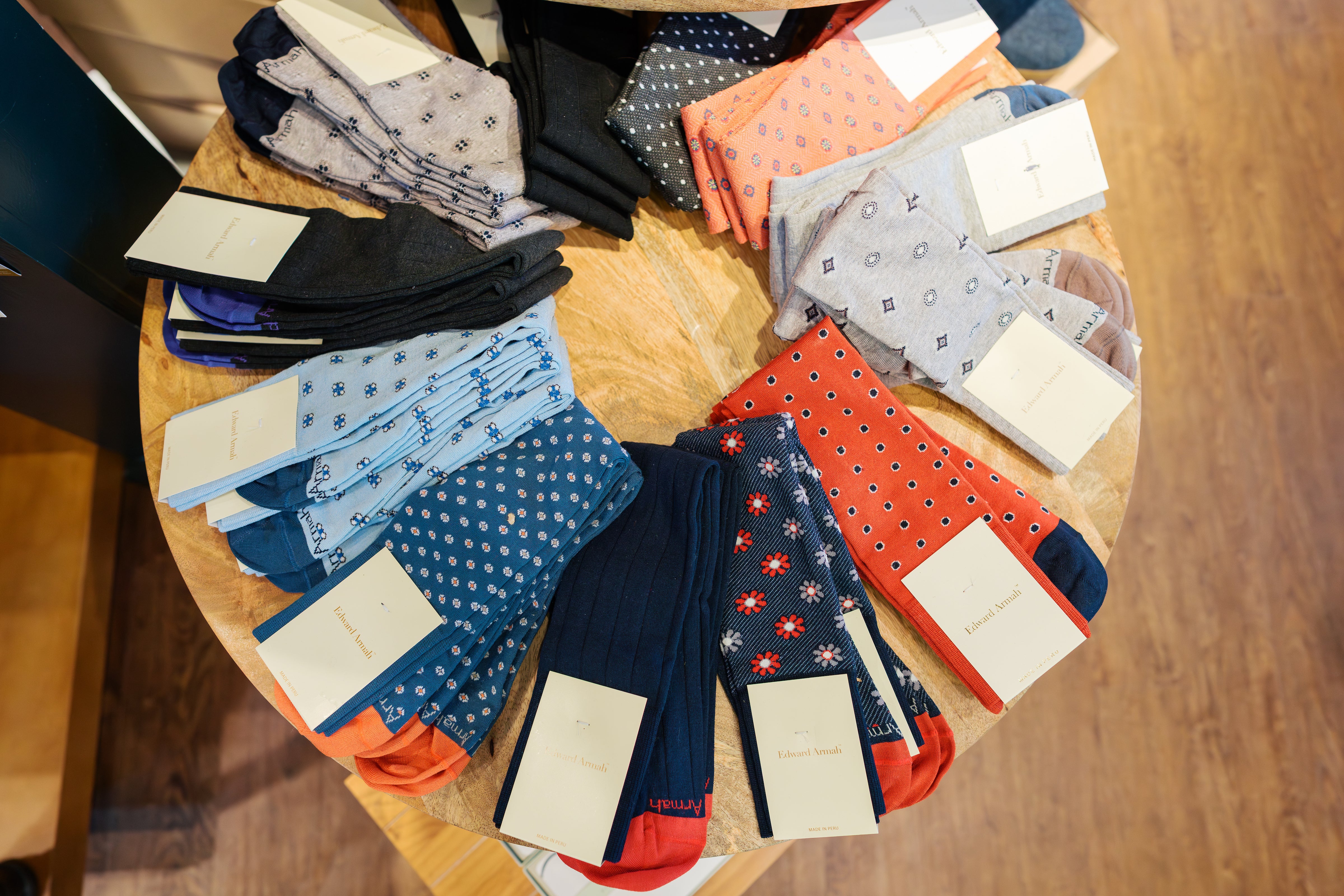 A round wooden table displays neatly arranged pairs of colorful patterned socks, each pair folded with a cream tag, showcasing various colors and designs, including blue, red, black, and gray tones.
