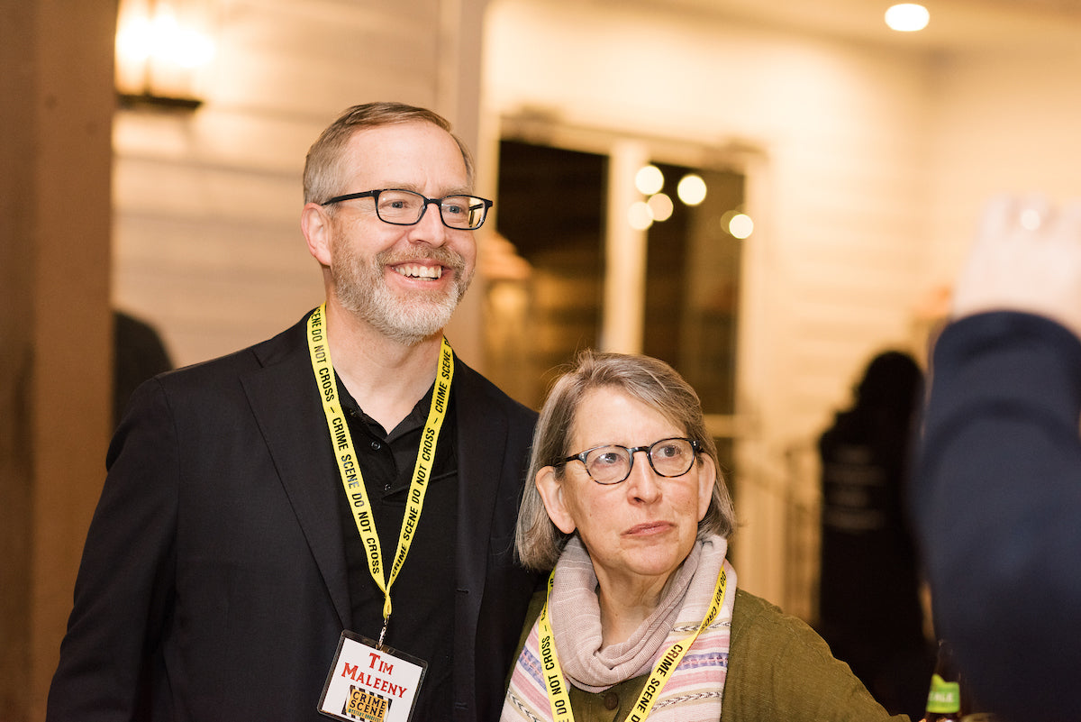 A man in a suit and a woman wearing glasses pose together, both smiling. They wear yellow “SHOW CREW” lanyards. The setting appears to be a warmly lit indoor-outdoor event.