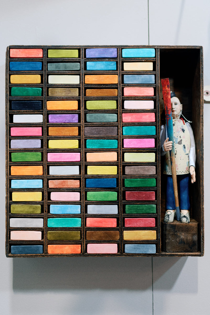 A wooden box holds rows of colorful pastel sticks, neatly organized by color. In the rightmost compartment stands a painted artist figurine holding a large red paintbrush.