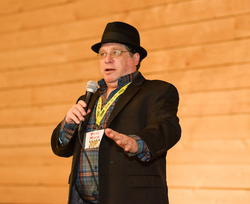 A man wearing a black hat, glasses, plaid shirt, and blazer holds a microphone and gestures while speaking. He wears a name tag that reads