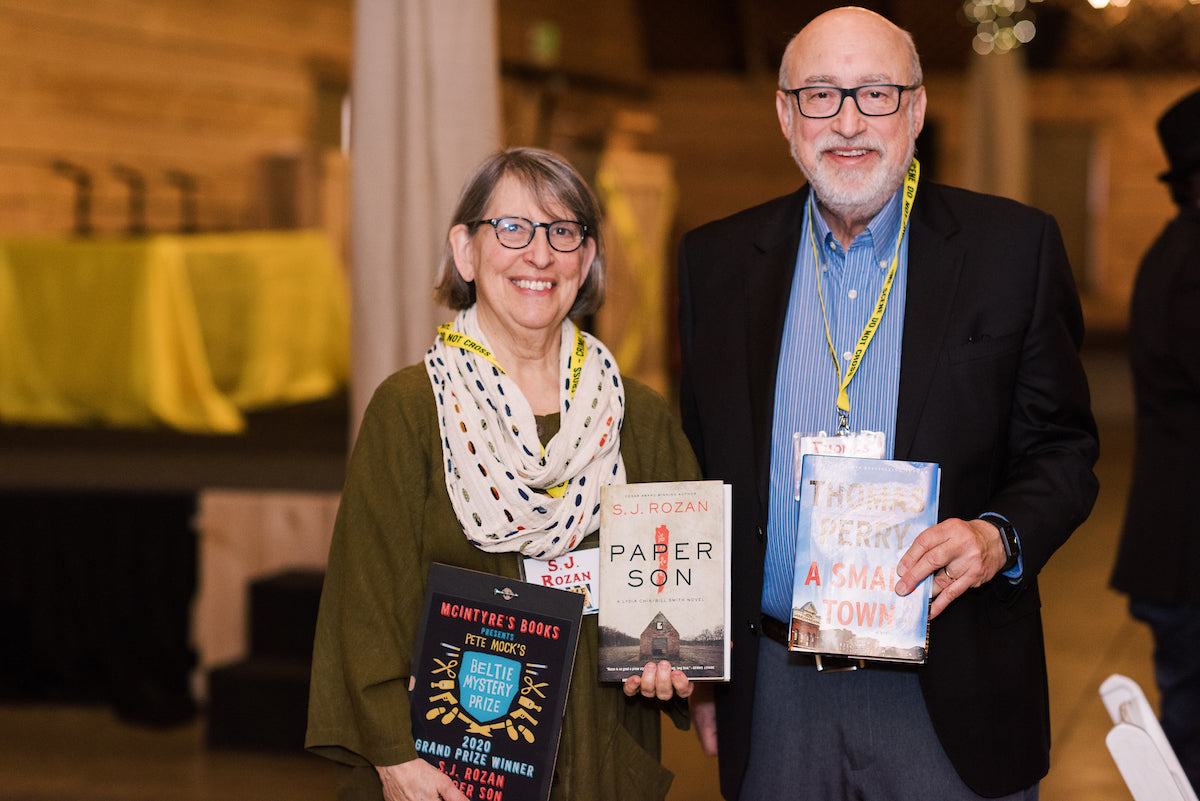 Two smiling older adults, a woman and a man, stand indoors holding books. Both wear glasses and yellow lanyards. The woman holds three books; the man holds two. The background is softly lit and out of focus.