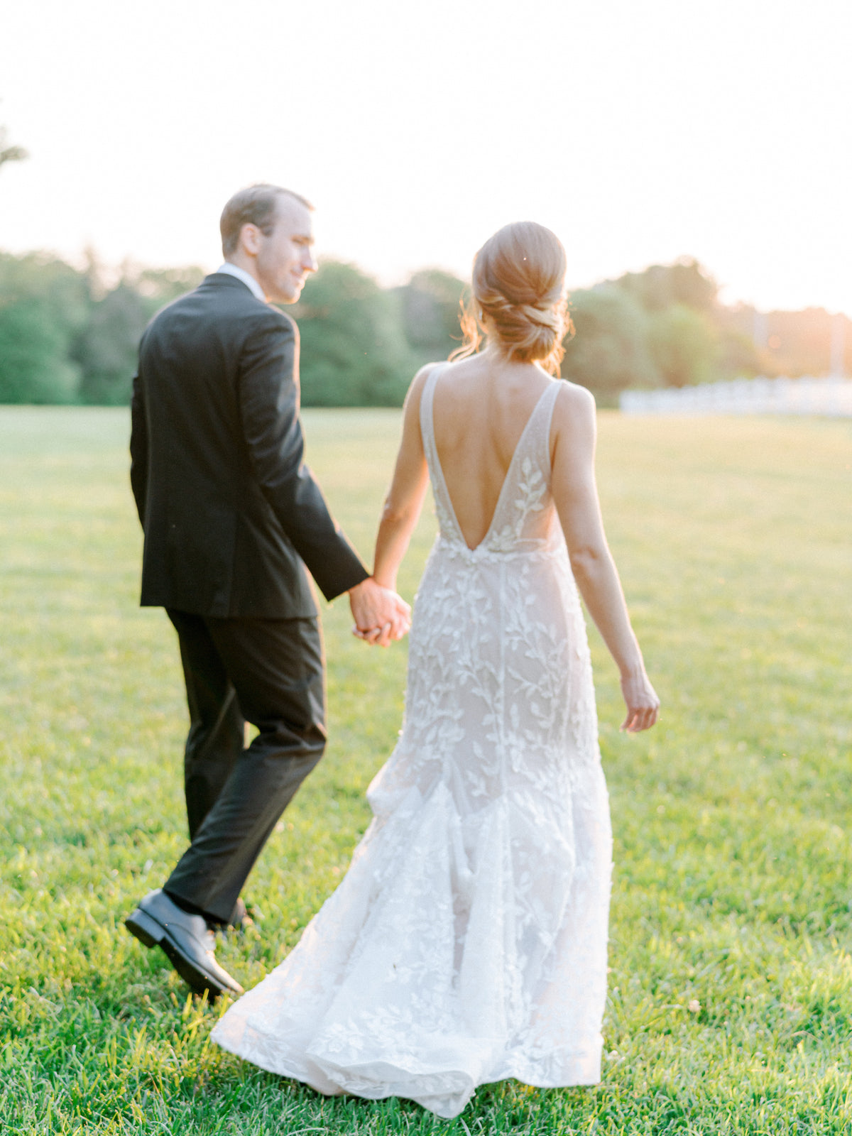 A bride in a lace wedding dress with a low back and a groom in a black suit walk hand in hand across a grassy field at sunset.
